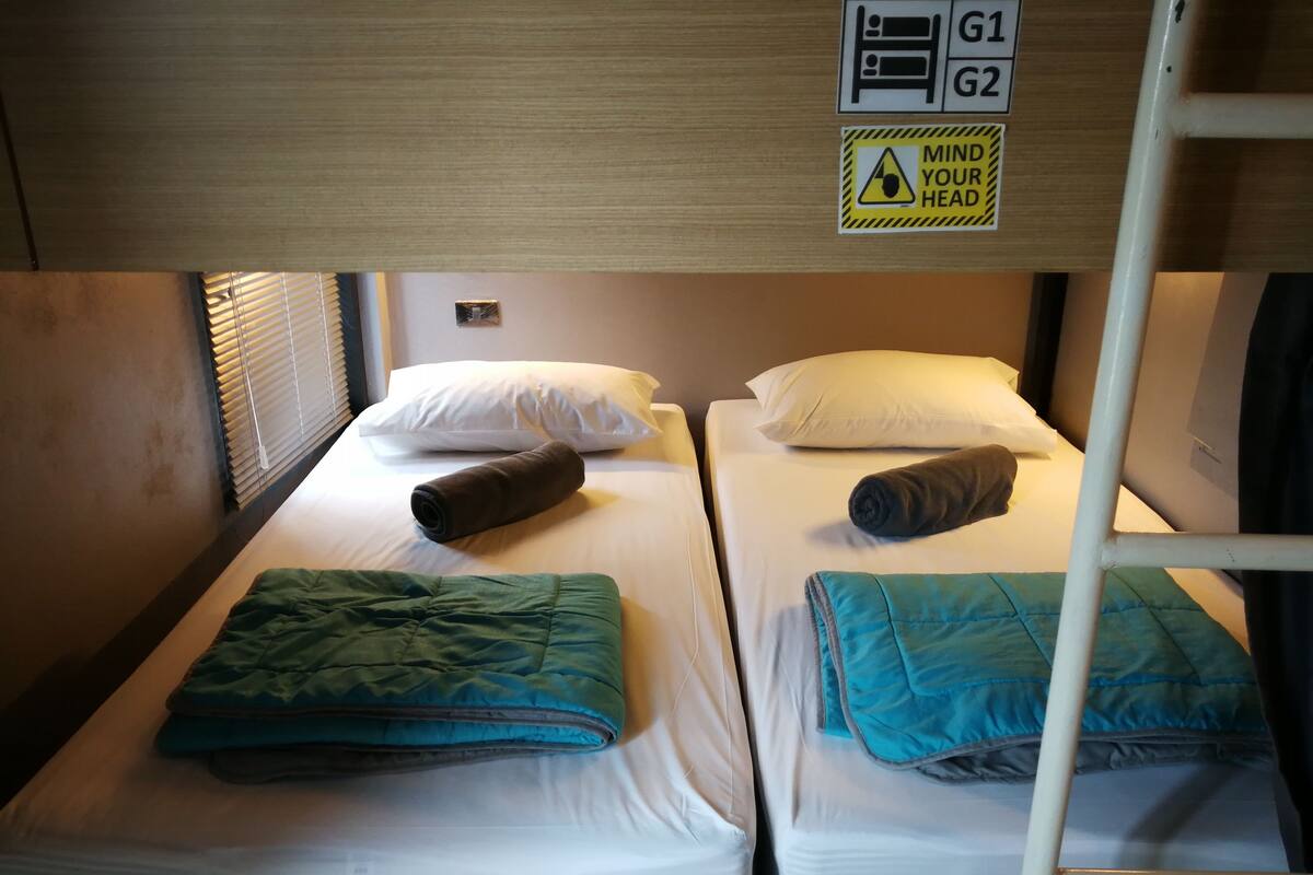 Habitación con literas, dos camas, cada una con una cobija enrollada y almohadas. Un letrero sobre las camas indica los números de habitación G1 y G2.