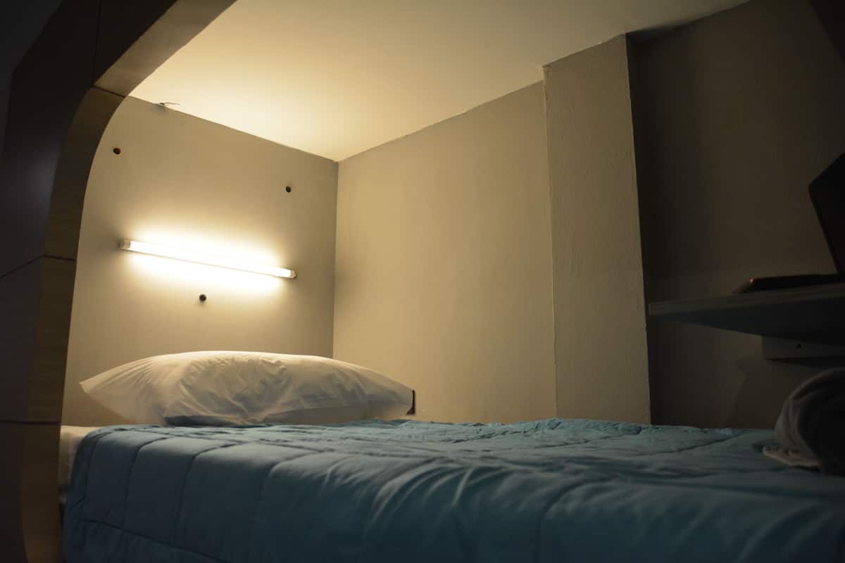Una habitación pequeña y tenue, con una cama individual, una lámpara en la pared y una vista parcial de una escalera.