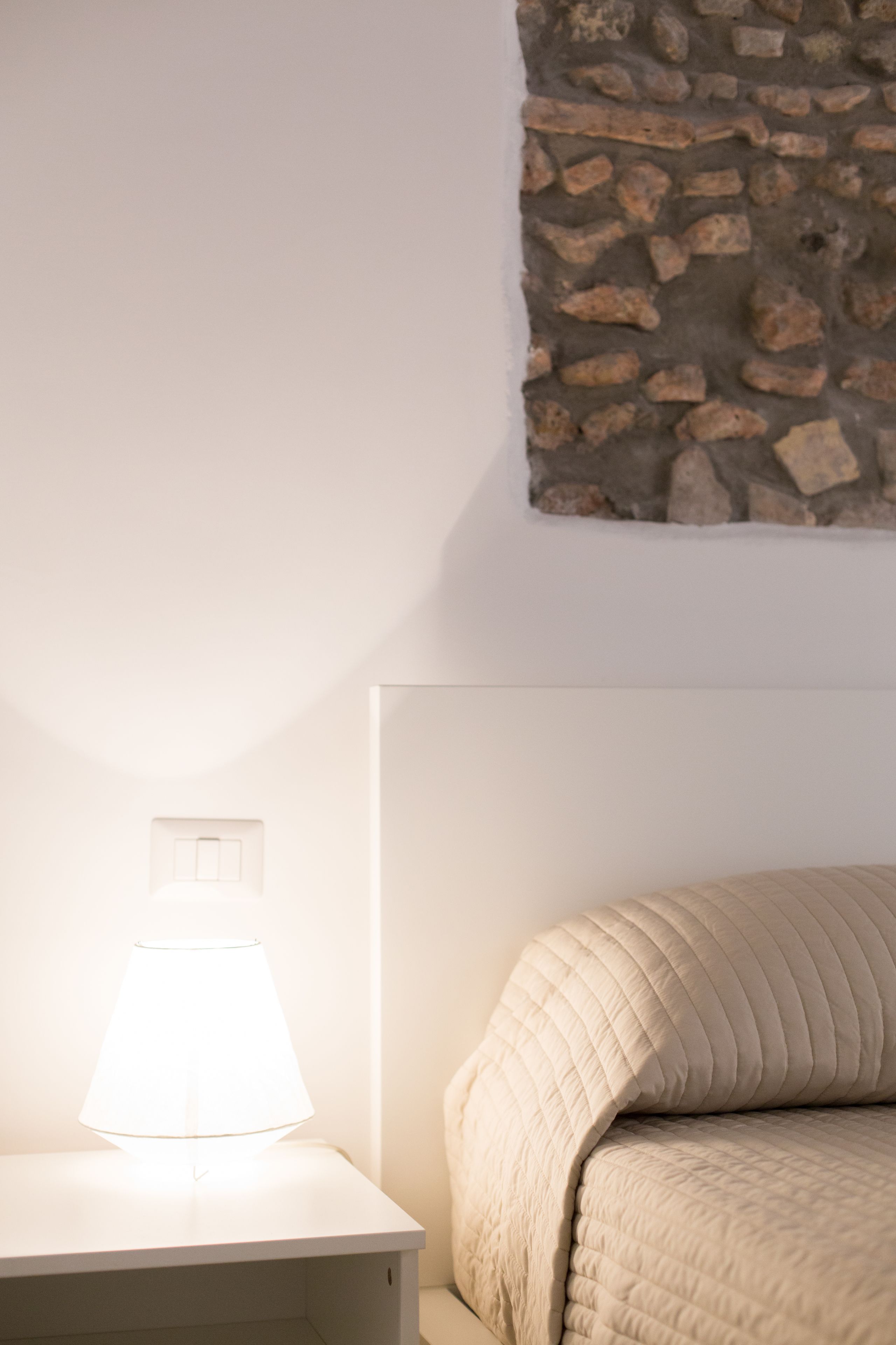 Een net opgemaakt bed met een wit hoofdeinde, een bedlampje en een stenen muur erboven.