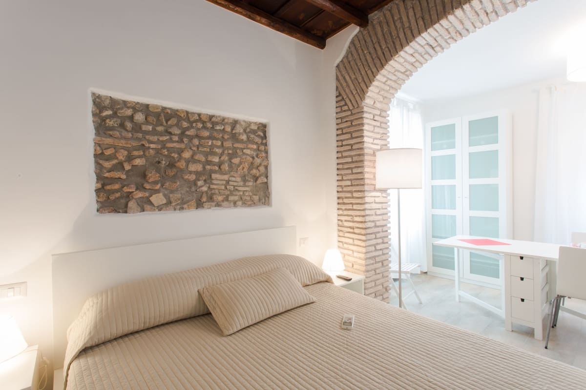 Ein Schlafzimmer mit Steinwand, einem Bett mit beige farbenem Bettzeug, einem Schreibtisch mit Lampe und einem Fenster mit satiniertem Glas.
