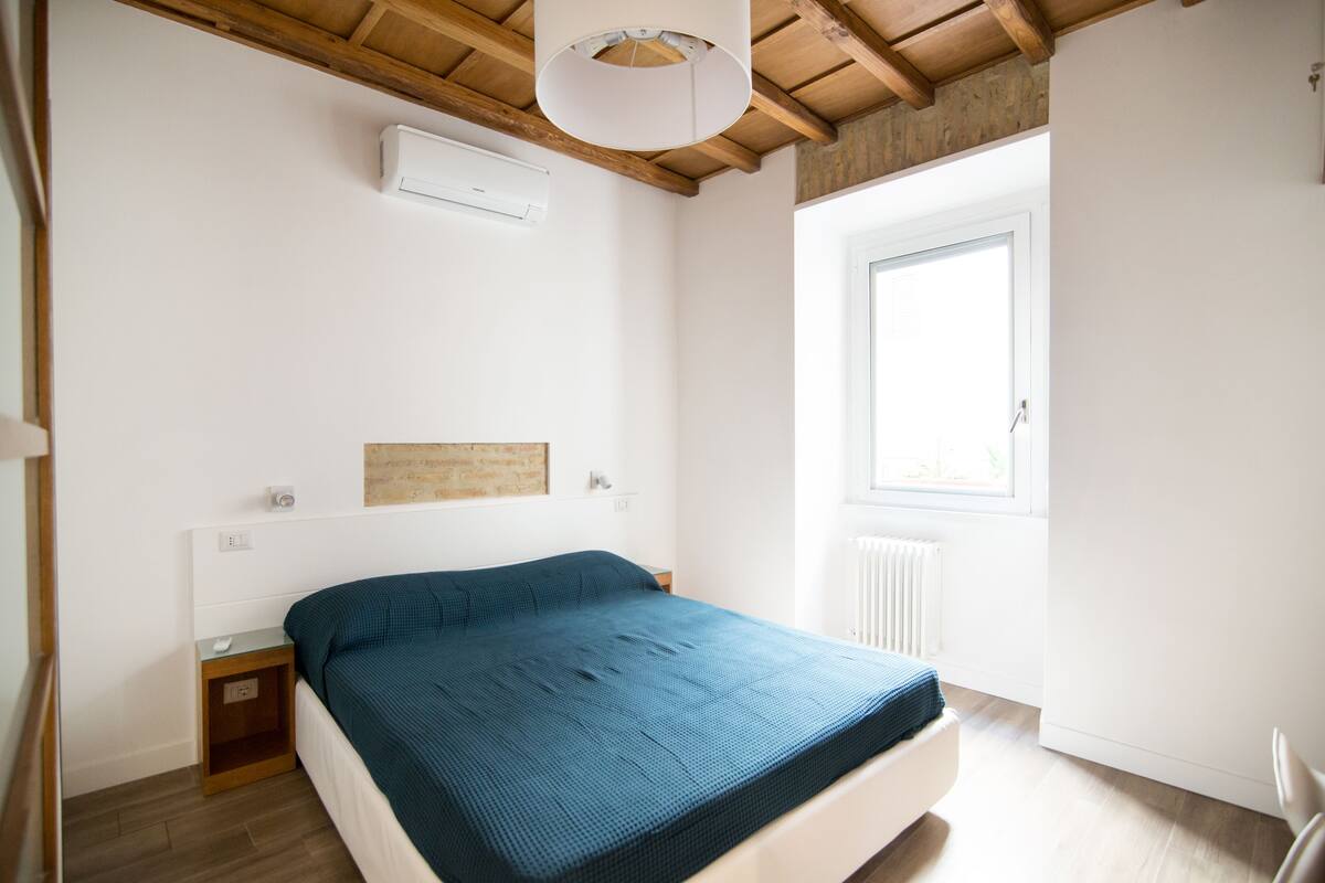 Ein Schlafzimmer mit Bett, Nachttisch, Deckenleuchte, Fenster und Heizkörper.