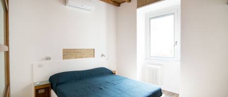 Een slaapkamer met een bed, nachtkastje, plafondlamp, raam en een radiator.