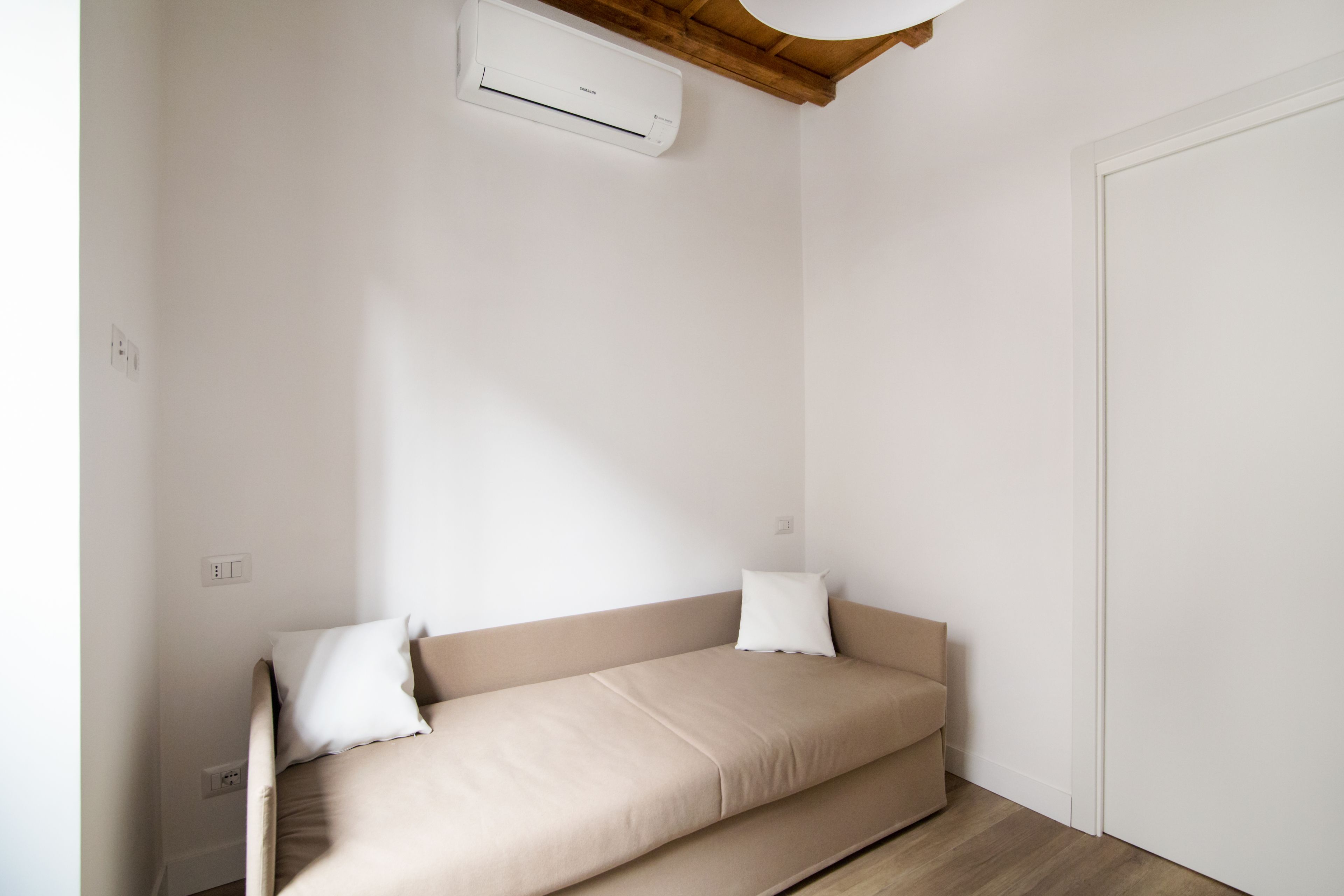 Ein kleines Zimmer mit einem Einzelbett, einer beigen Matratze, weißen Kissen, einer Holzbalkendecke und einer weißen Wand.