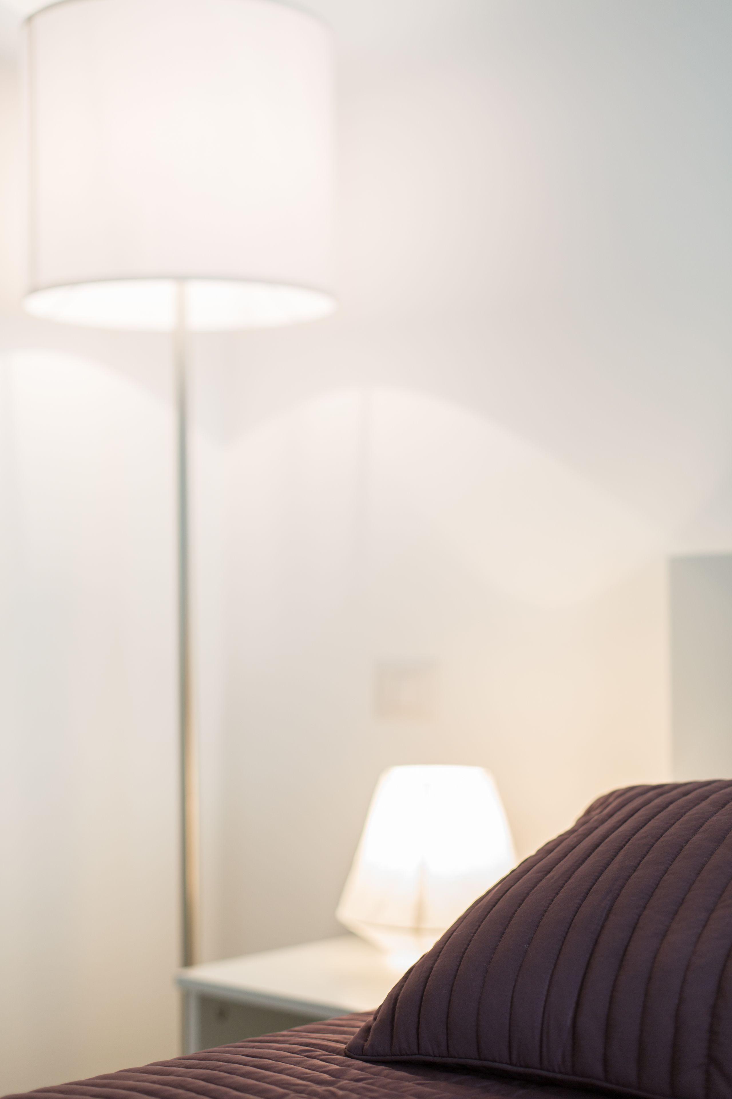 Una cama con cabecera oscura, una lámpara de noche y una lámpara de pie en una habitación con pared blanca.