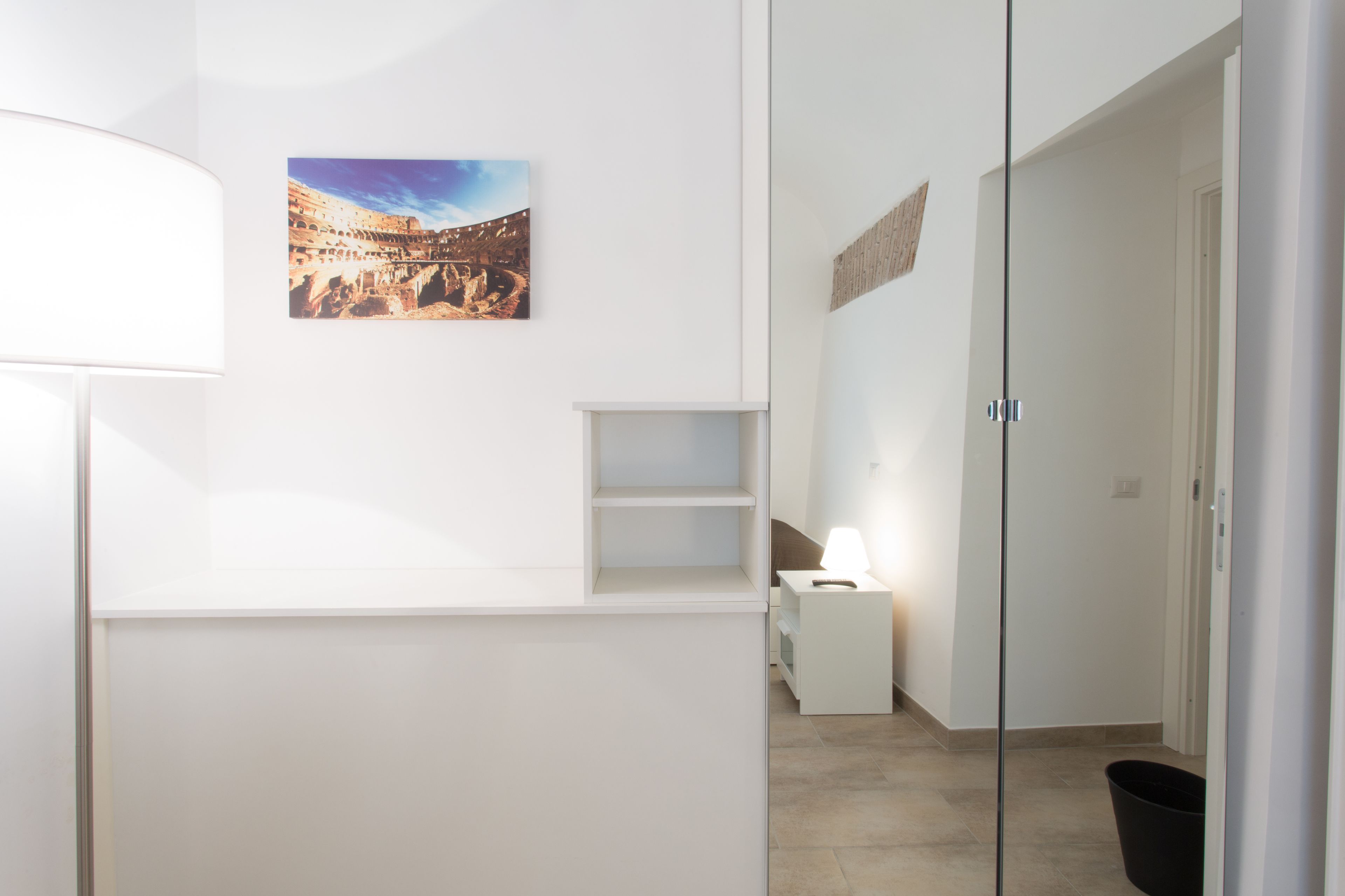 A modern hotel room with a white reception desk, a small bedside table with a lamp, a picture of a historical site on the wall, and a glass door leading to another room.