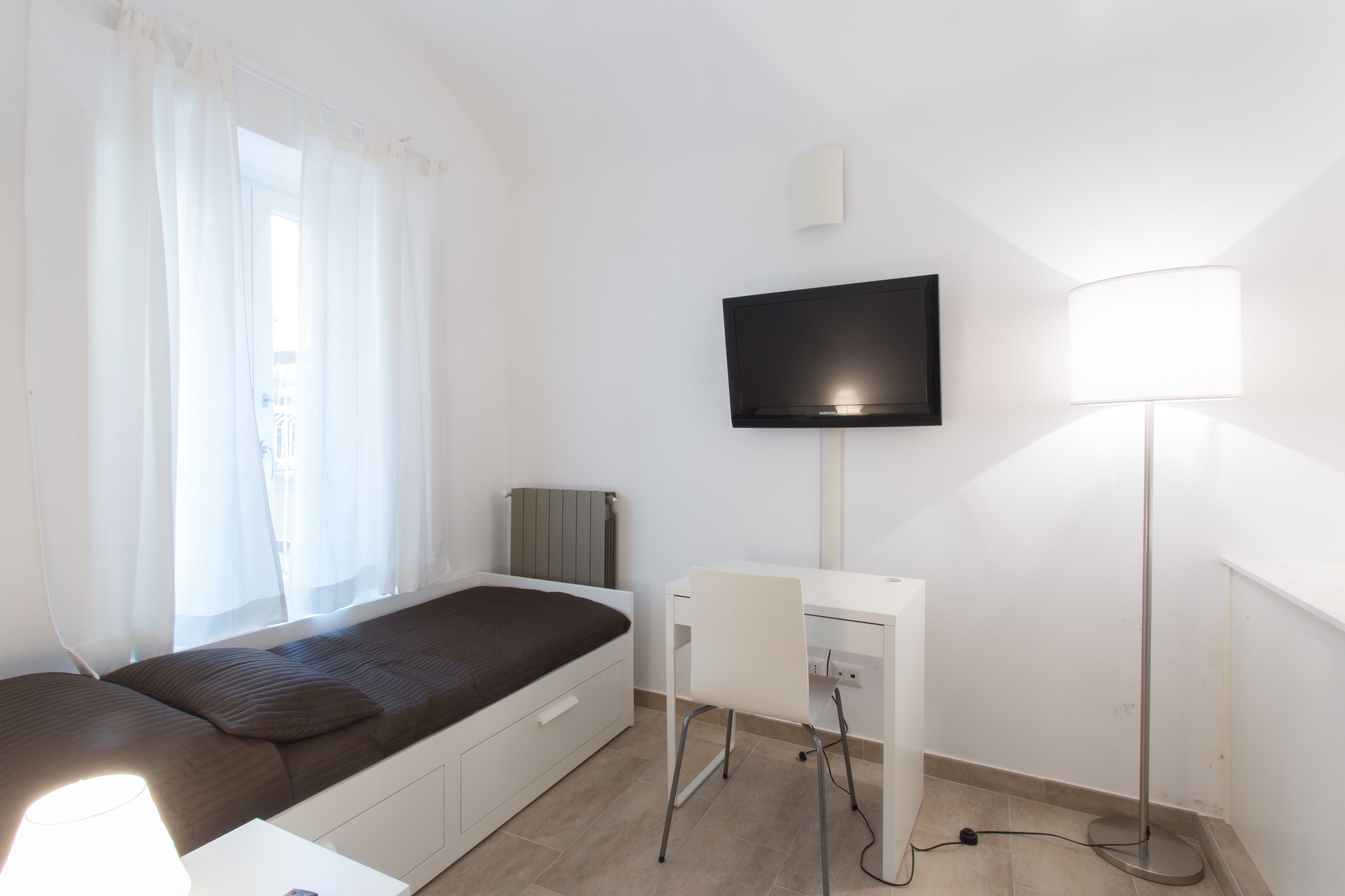 A bedroom with a bed, a desk, a chair, a TV, and a lamp.