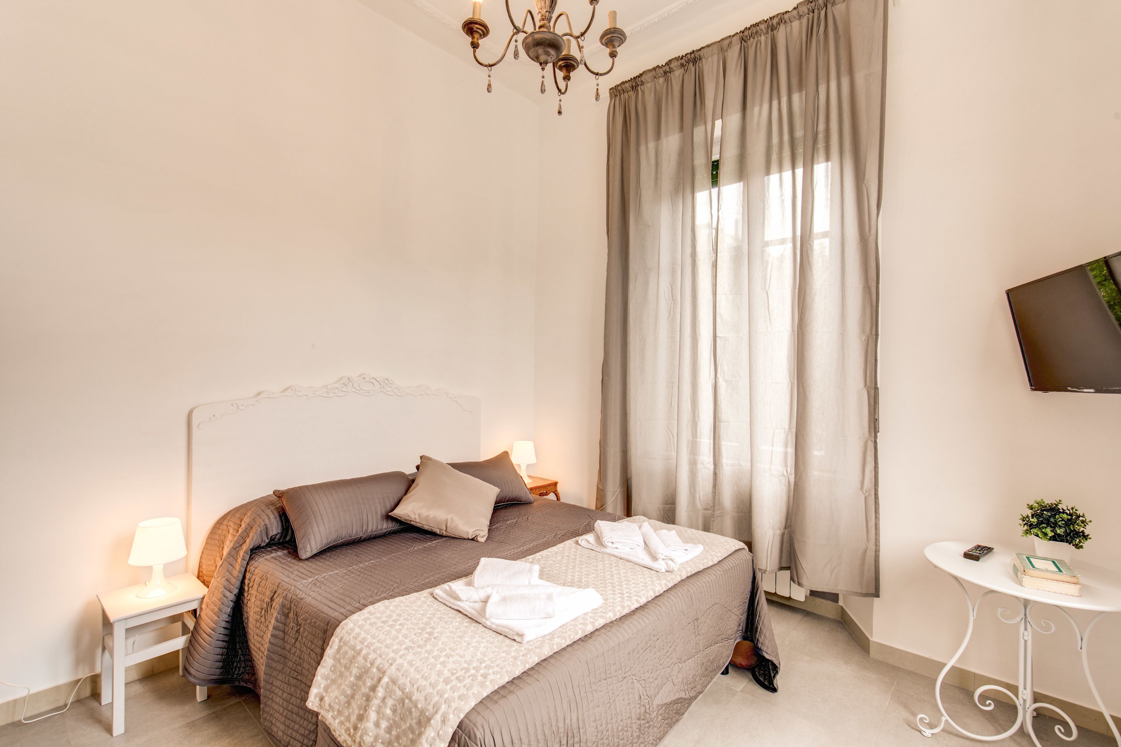 A bedroom with a large bed, a bedside table with a lamp, a small table with a plant, and a TV on the wall.