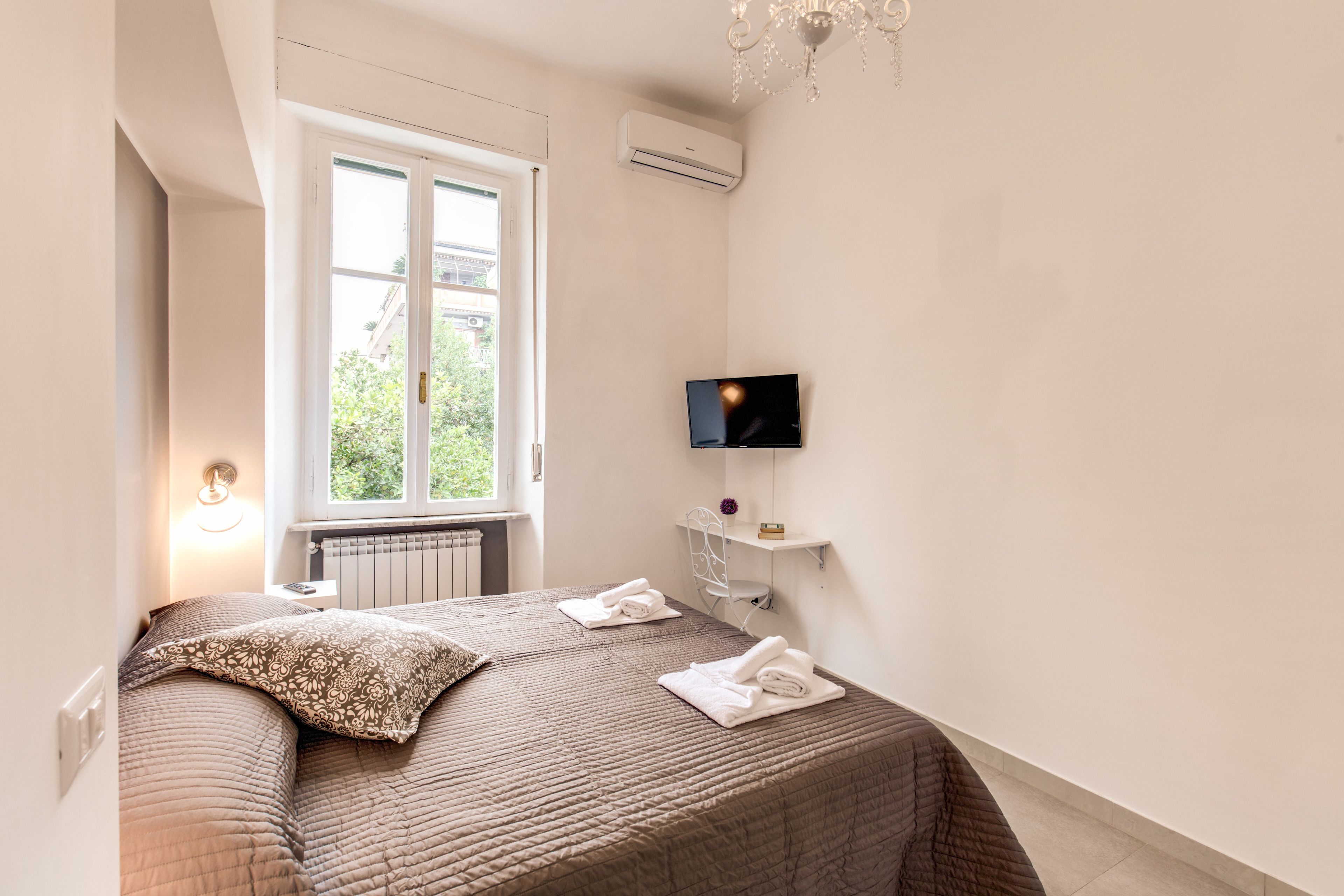 A bedroom with a bed, a TV mounted on the wall, a window, and an air conditioning unit.