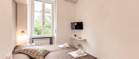 A bedroom with a bed, a TV mounted on the wall, a window, and an air conditioning unit.