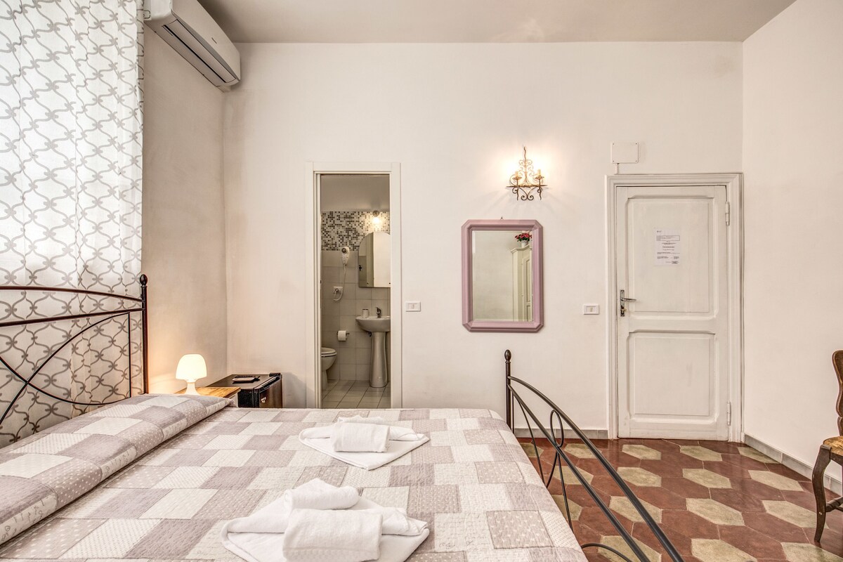 A bedroom with a bed, a bedside table, a mirror, a door, and a bathroom visible through an open door.