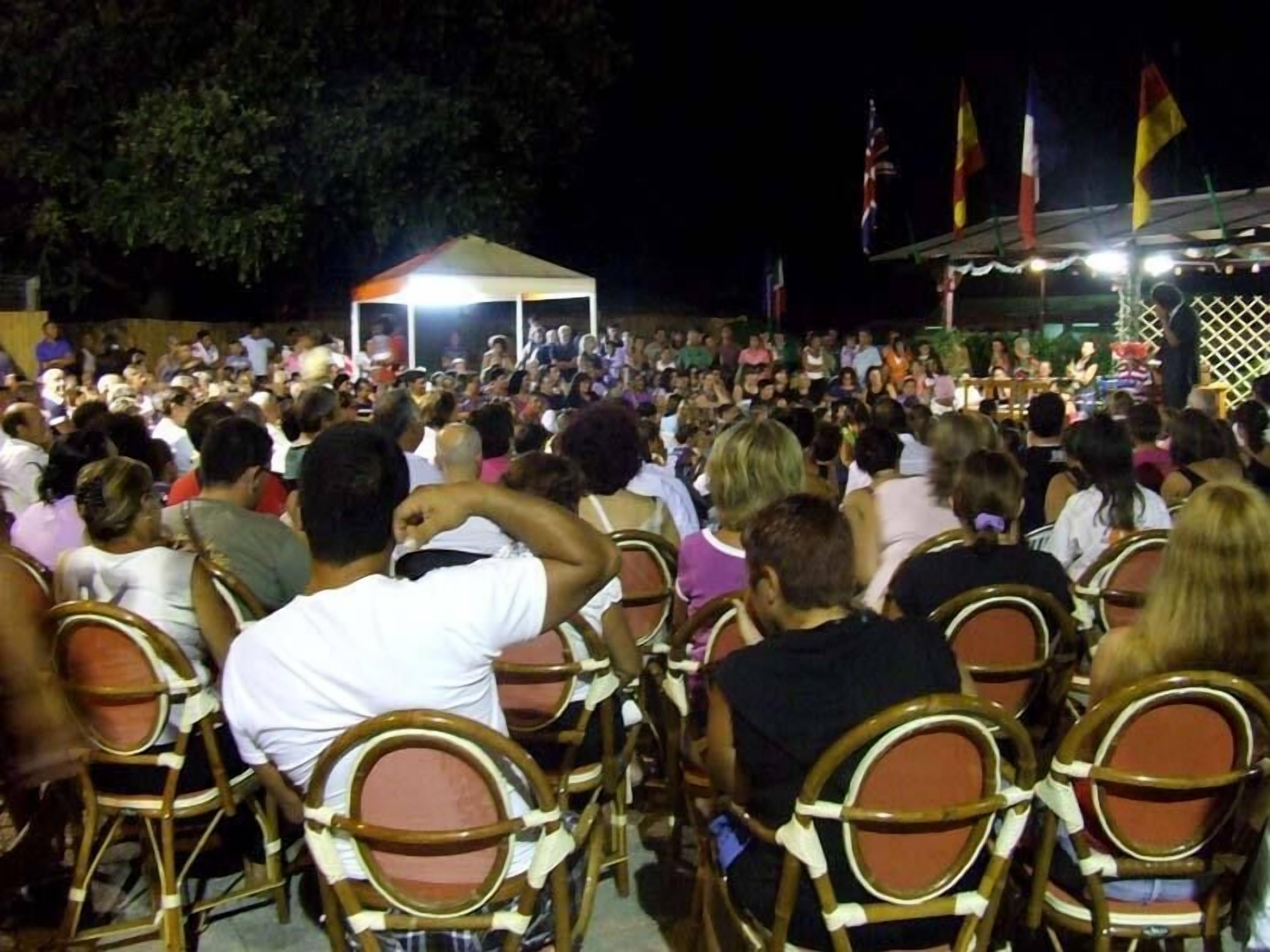 A crowded outdoor event with people seated on wooden chairs facing a stage.
