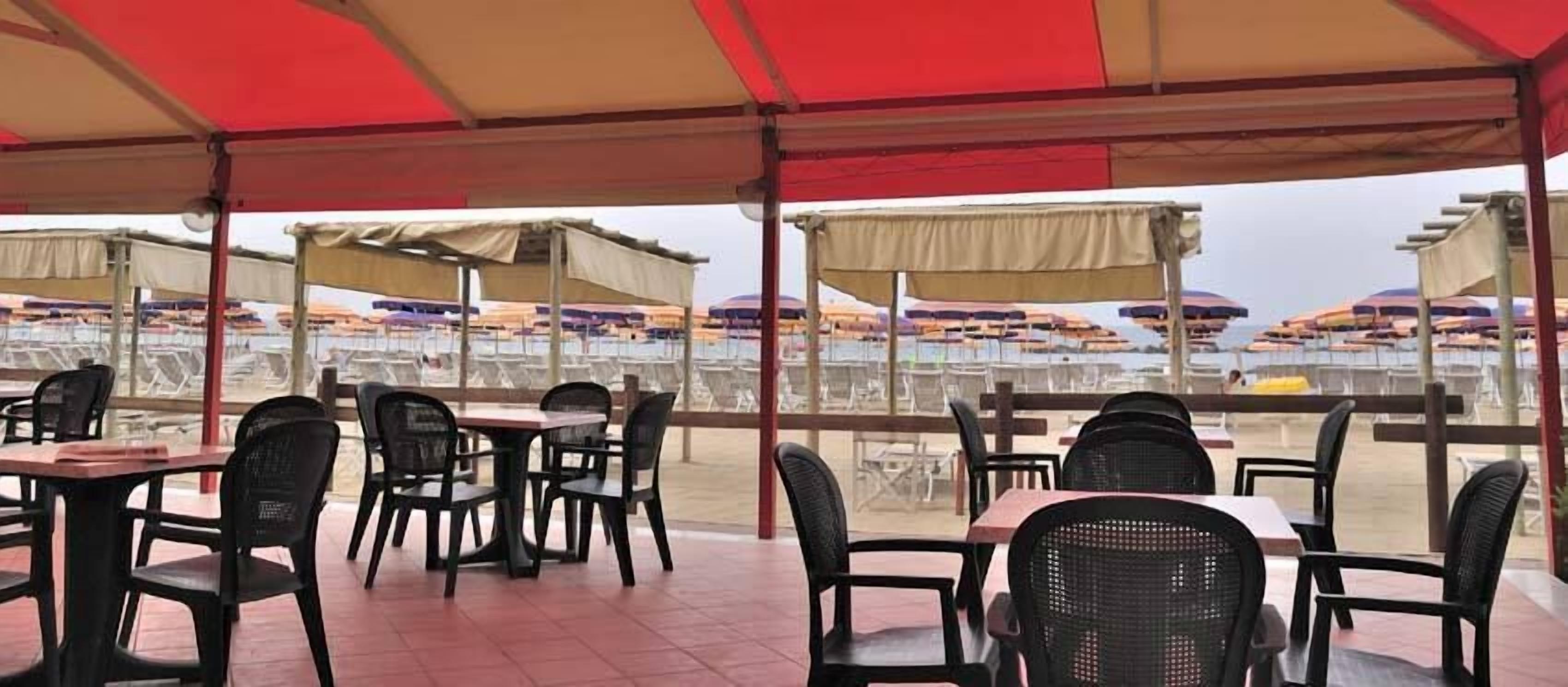 Area pranzo all'aperto con tavoli e sedie sotto una tettoia, con vista su una spiaggia con ombrelloni.