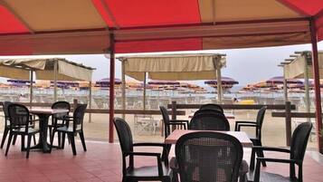 Area pranzo all'aperto con tavoli e sedie sotto una tettoia, con vista su una spiaggia con ombrelloni.