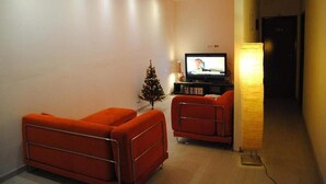 Ein modernes Wohnzimmer mit zwei orangefarbenen Sesseln, einer kleinen Weihnachtstanne, einem Fernseher und einer Stehlampe.