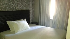 Ein ordentlich bezogenes Bett mit weißem Kopfkissen und dunklem Kopfteil in einem Zimmer mit raumhoher, gemusterter Tapete und einem Fenster mit Vorhängen.