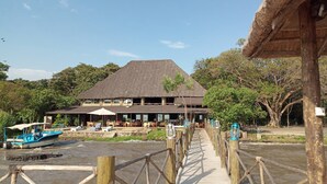 Um edifício com telhado de palha, mesas ao ar livre e uma passarela de madeira sobre a água.