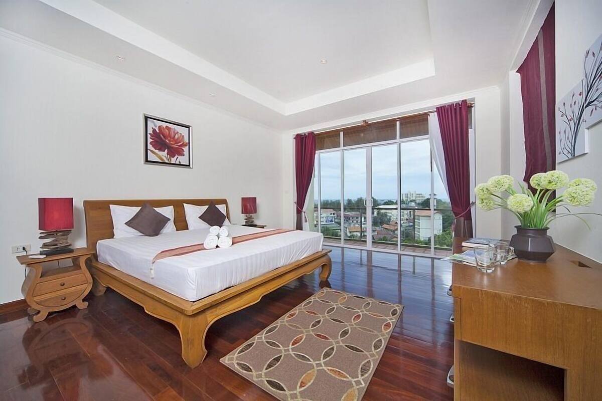 Quarto com cama grande, cabeceira de madeira, mesa de cabeceira, escrivaninha, janela com cortinas e vista para a cidade.