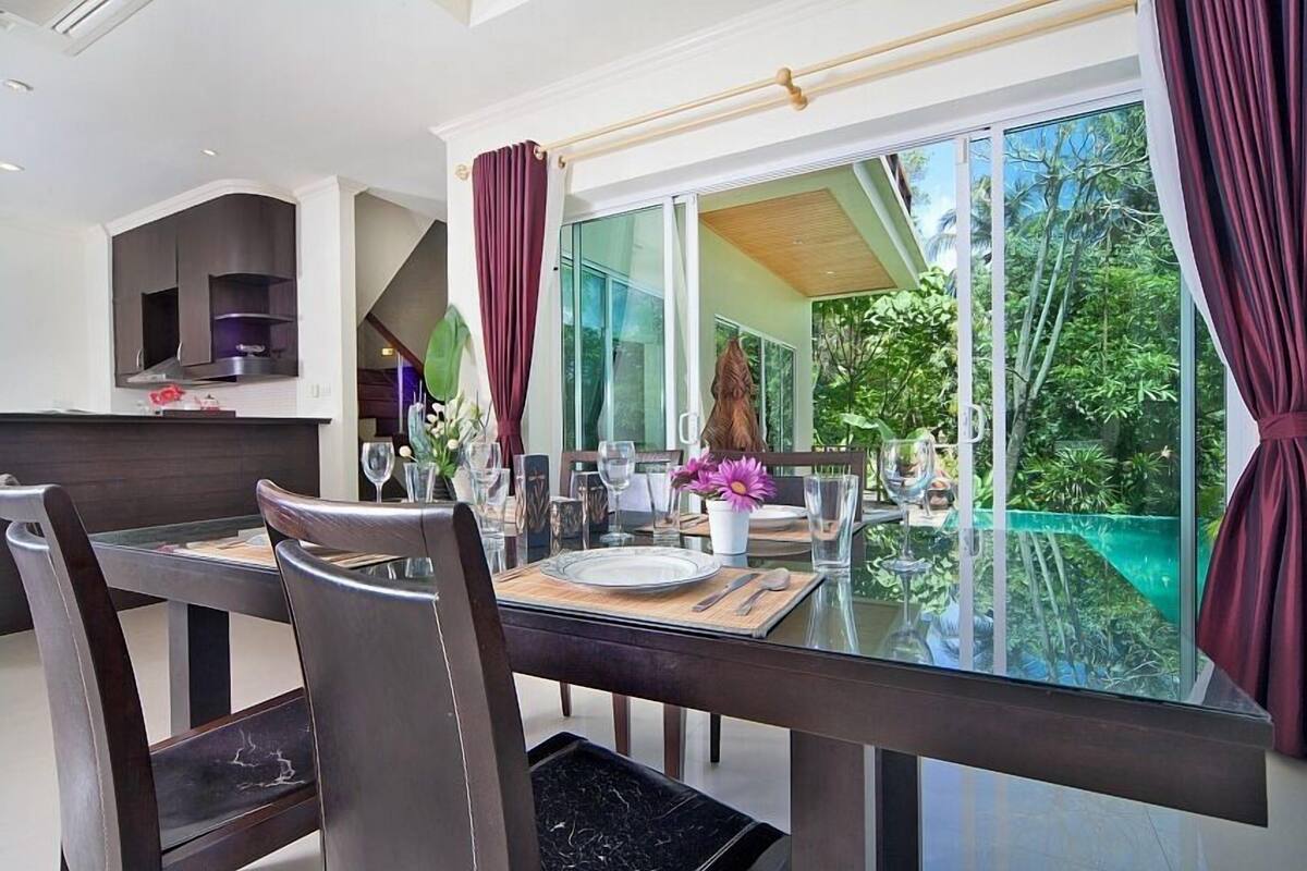 Uma sala de jantar moderna com mesa e cadeiras de vidro, vista para uma piscina e uma cozinha ao fundo.
