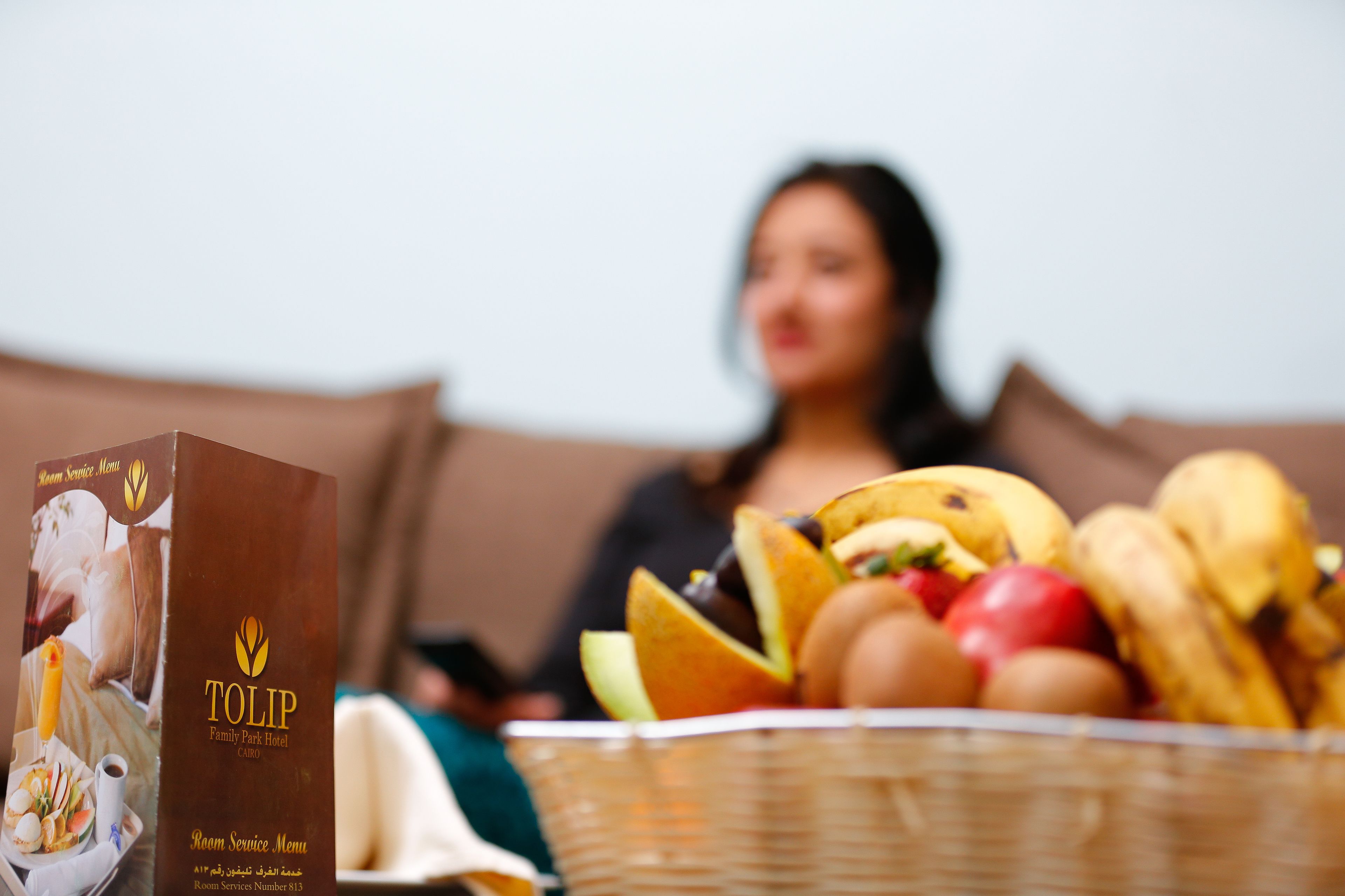 A room service menu box for the Tulip Family Park Hotel, a basket of fruits and bread, and a blurred figure in the background.