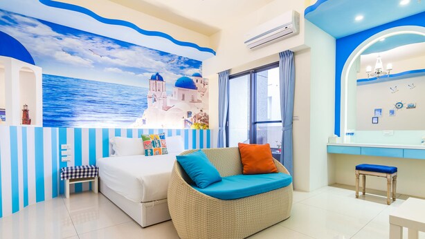 Quarto com cama, sofá de vime com almofadas, um pequeno banquinho, toucador com espelho e um mural na parede com tema costeiro.