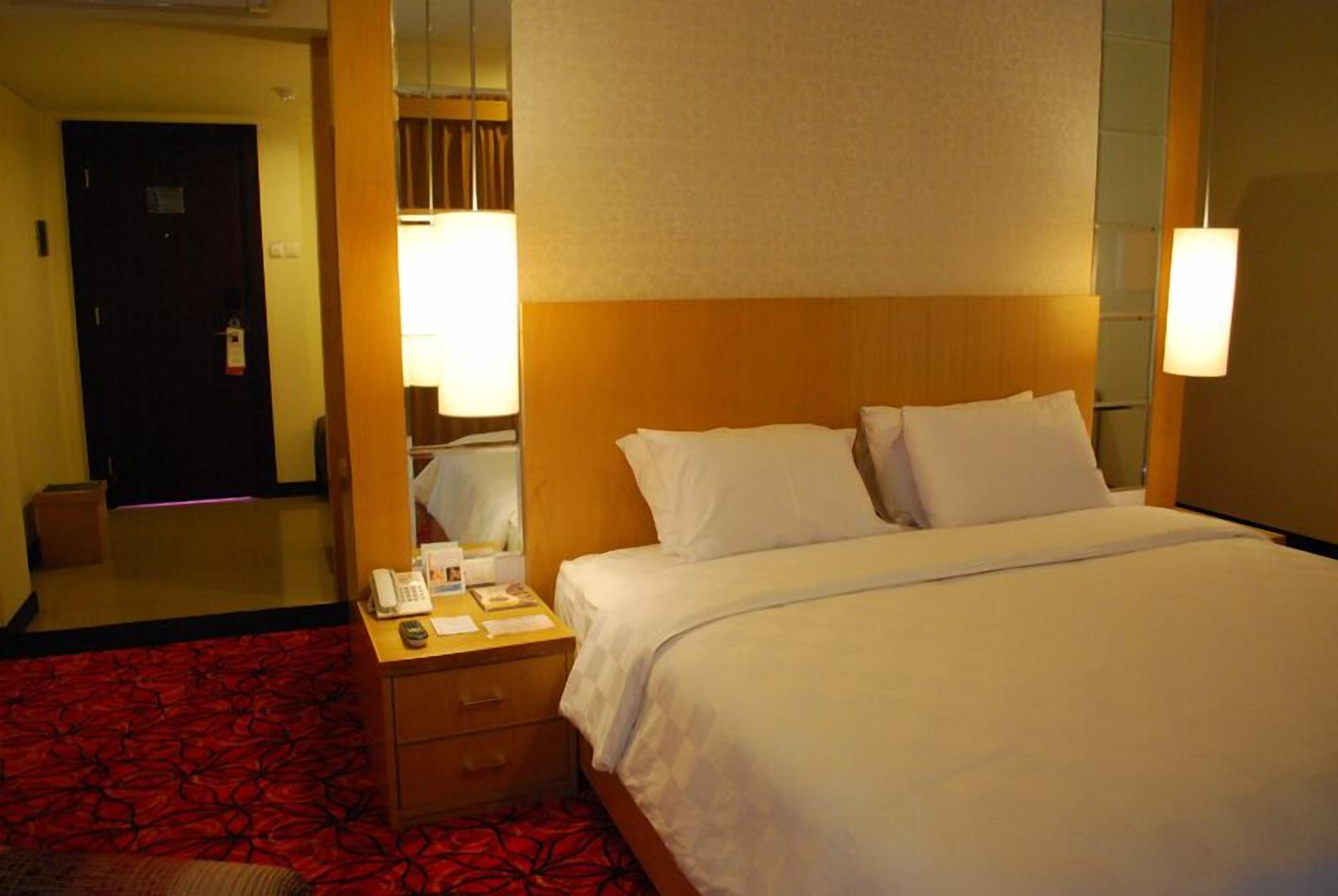 A hotel room with a bed, a bedside table with a lamp, a mirror, and a red patterned carpet.