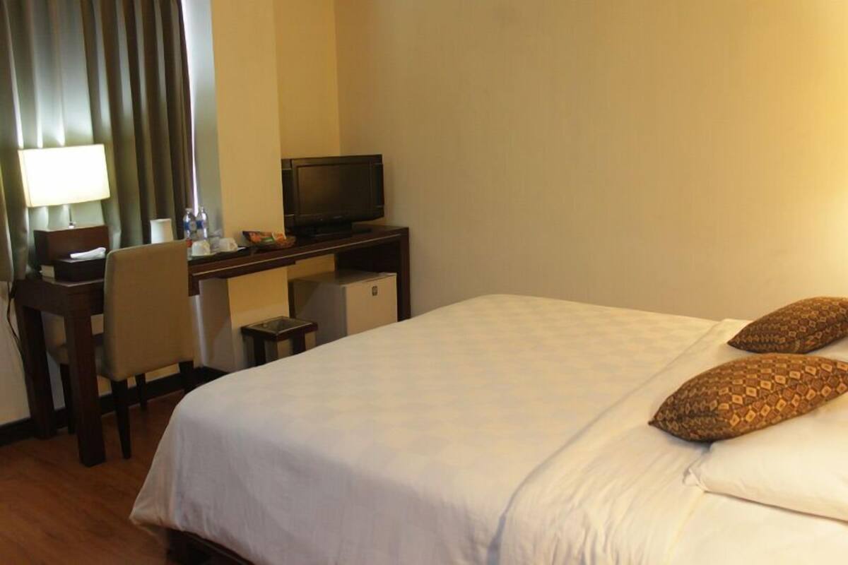 A hotel room with a bed, a desk with a television, a chair, and a lamp.