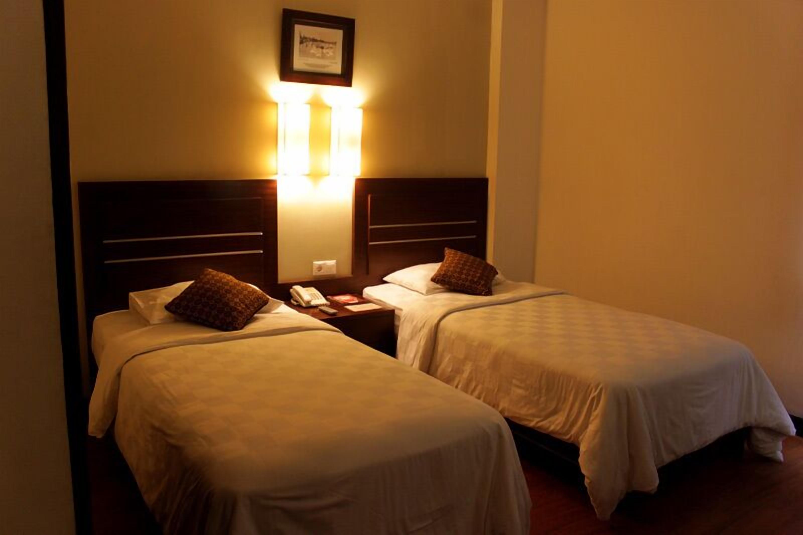 A hotel room with two single beds, a nightstand with a phone, and a wall-mounted light fixture.