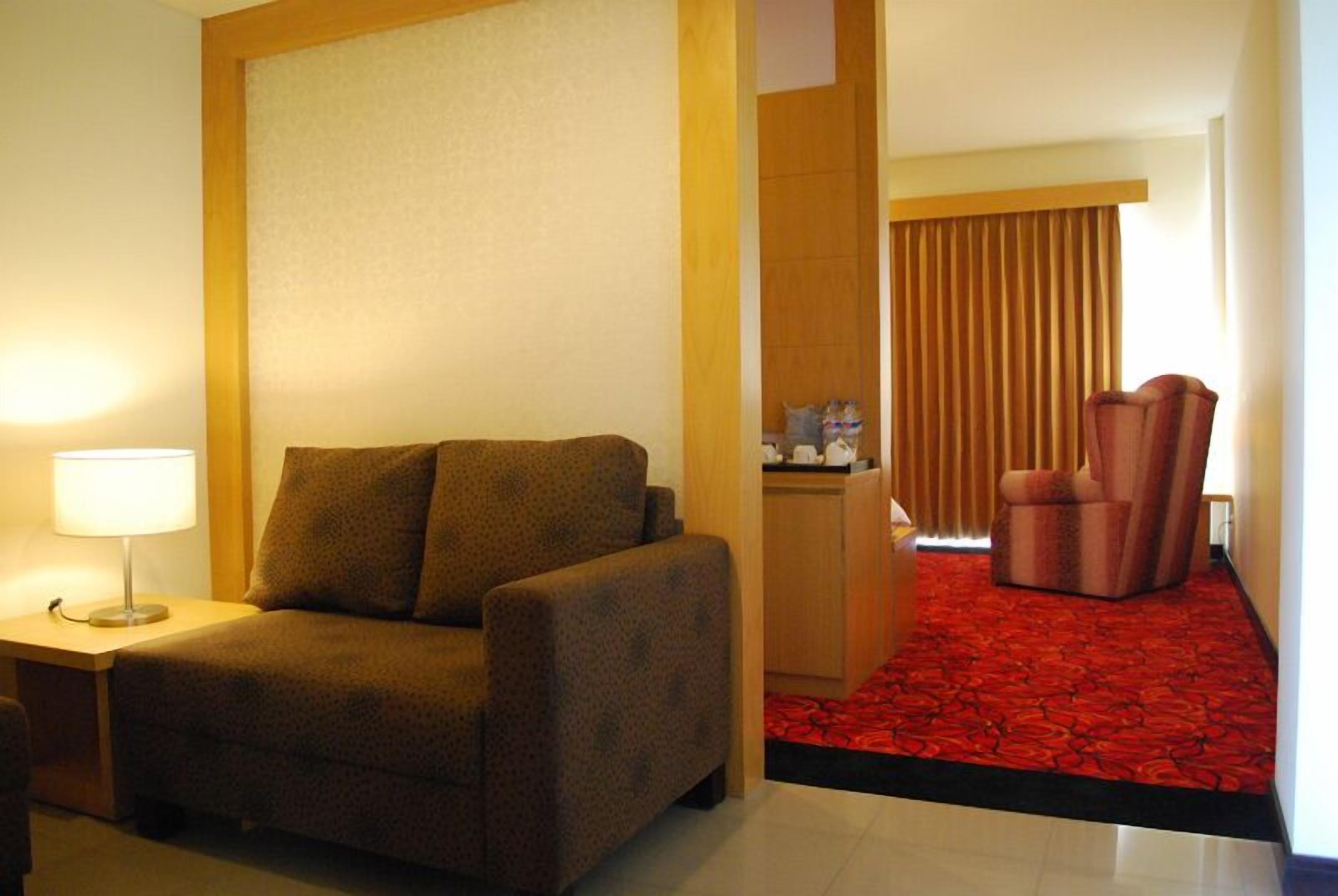 Habitación de hotel con sofá, escritorio con lámpara, aparador de madera y zona con alfombra roja.