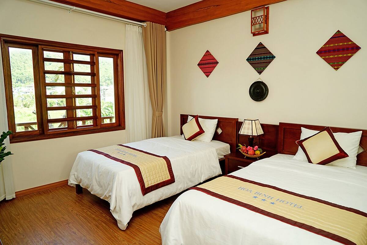 Quarto de hotel com duas camas, soalho de madeira, uma janela grande com cortinas e detalhes decorativos nas paredes.