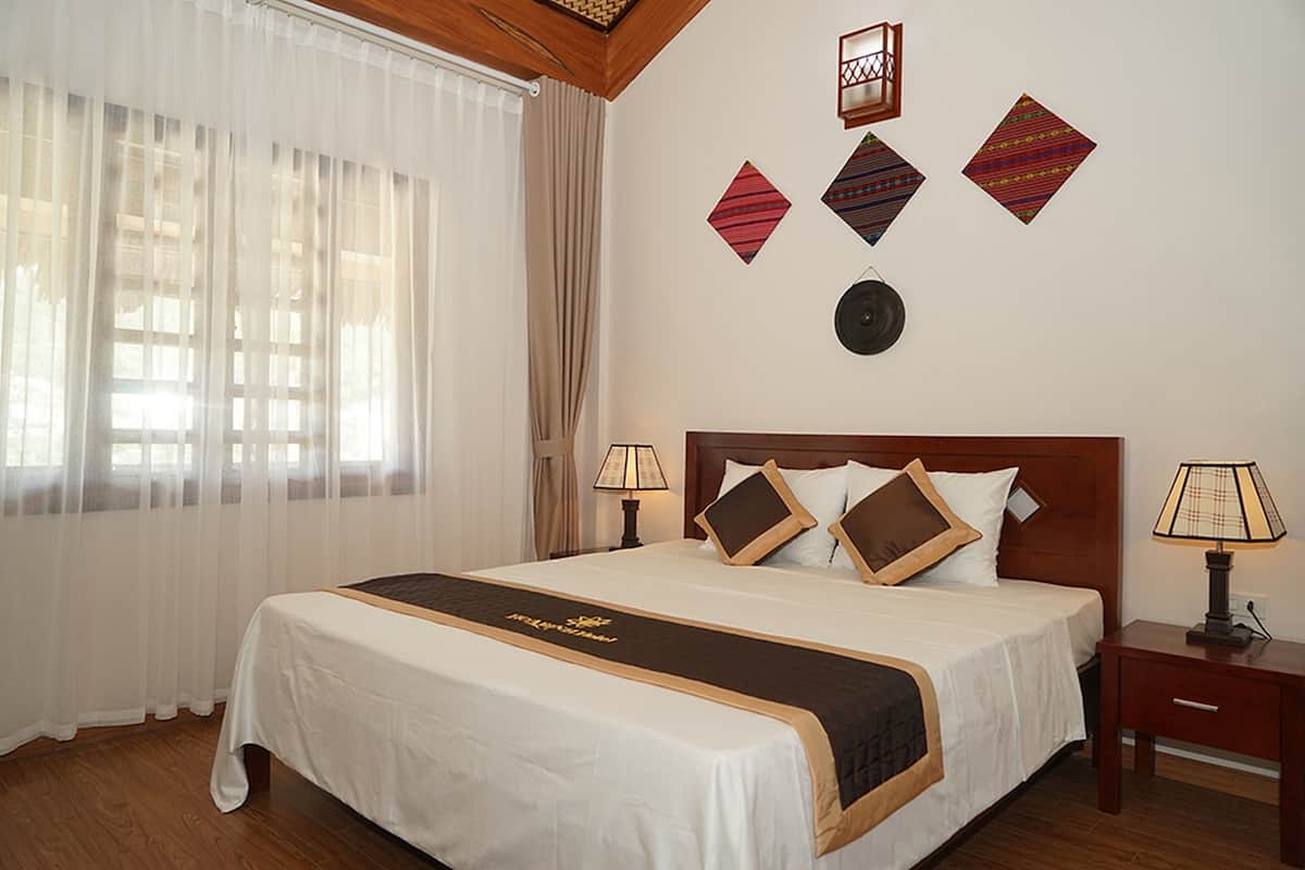 Cama bem arrumada com colcha branca e marrom, duas luminárias de cabeceira e uma cabeceira de madeira.
