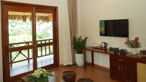 Une pièce avec un plancher en bois, un téléviseur fixé au mur, une plante en pot, une table avec une bouilloire et une grande fenêtre donnant sur un paysage verdoyant.