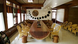 Une salle de conférence avec une longue table en bois entourée de chaises en osier, un mur décoratif orné d’assiettes et une bibliothèque remplie de magazines.