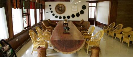 Un salón de conferencias con una larga mesa de madera rodeada de sillas de mimbre, una pared decorativa con platos y una estantería con revistas.