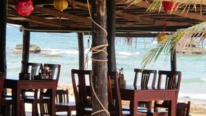 An outdoor wooden table and chairs set up under a thatched roof, overlooking a beach with calm waters and rocks. - Nhat Lan Resort (Phu Quoc)