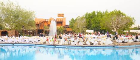 Una recepción de boda junto a la piscina, con invitados sentados en mesas, una fuente y un edificio al fondo.