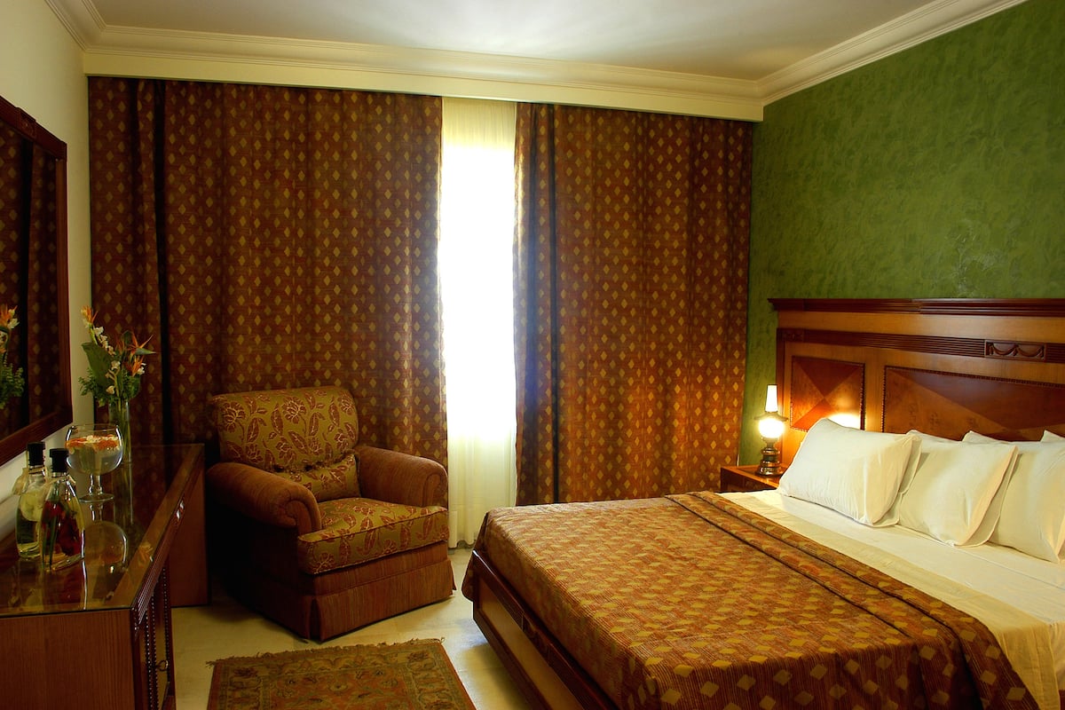 A hotel room with a bed, a chair, a nightstand with a lamp, and a desk with a vase and bottles.