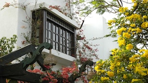 Un edificio tradicional con fachada blanca, una ventana con contraventanas de madera oscura y un balcón con macetas colgantes.