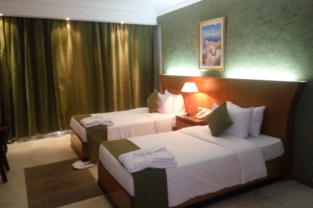 A hotel room with two beds, a nightstand with a lamp, a painting on the wall, and a green curtain.