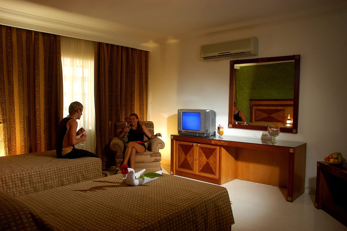 A hotel room with two beds, a television, a mirror, and a small table with fruit.