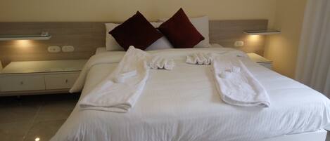 Una cama bien hecha con sábanas blancas y dos almohadas rojas, una mesita de noche con lámpara, y un marco de foto colgado en la pared.