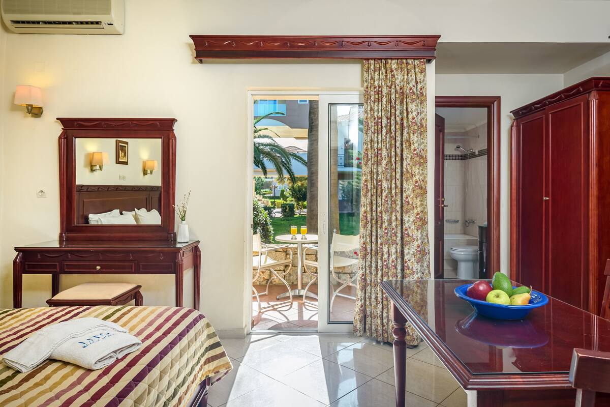 A hotel room with a bed, a desk, a mirror, a bathroom visible through a glass door, and a balcony with outdoor furniture.