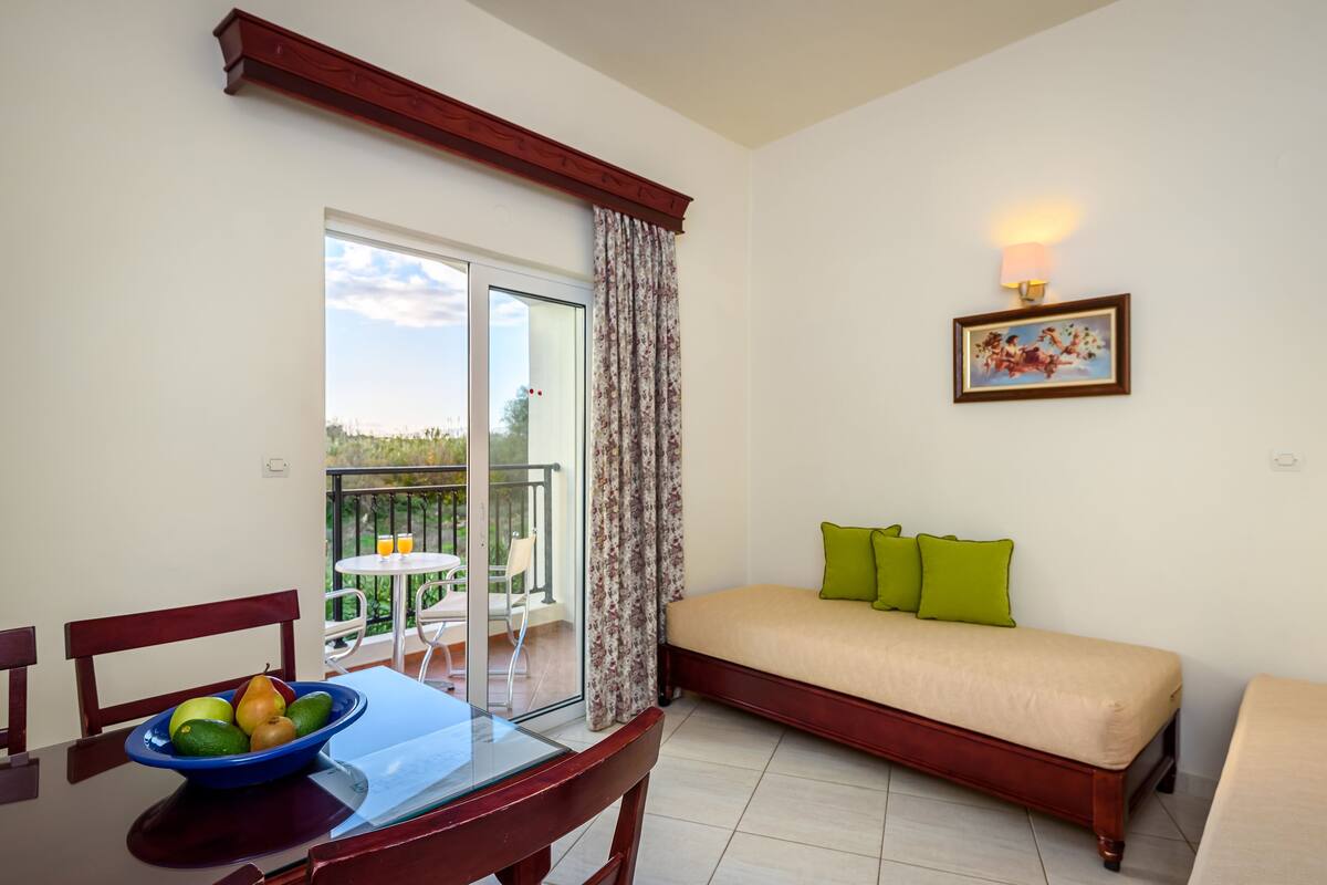 A room with a dining table, a bowl of fruit, a sofa with green cushions, a painting on the wall, and a balcony with a chair and table.