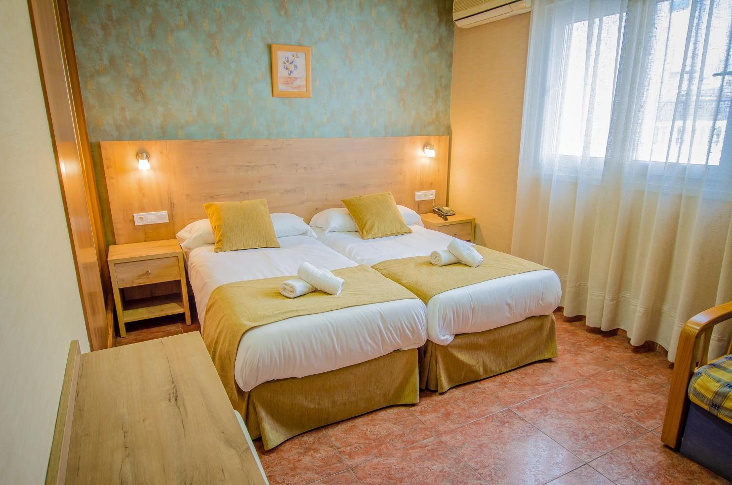 A double room with two beds, each with a headboard and bedside tables. There is a small table in the corner and a window with curtains.