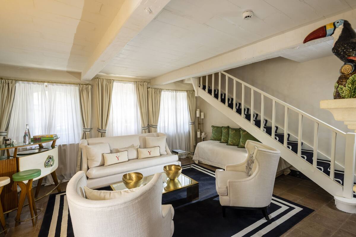 Un salon spacieux comprenant un canapé, des fauteuils et une table basse. On peut apercevoir un escalier orné d’une sculpture représentant un oiseau.