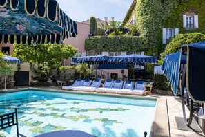 Un espace piscine doté de chaises longues et de parasols, entouré d’une végétation luxuriante et bordé d’un bâtiment dont les murs sont recouverts de lierre.