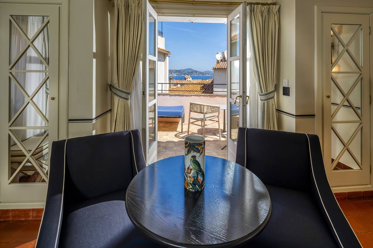 Une pièce avec une table ronde et des chaises, un vase décoratif, et une vue sur un balcon avec une table et des chaises à l’extérieur.