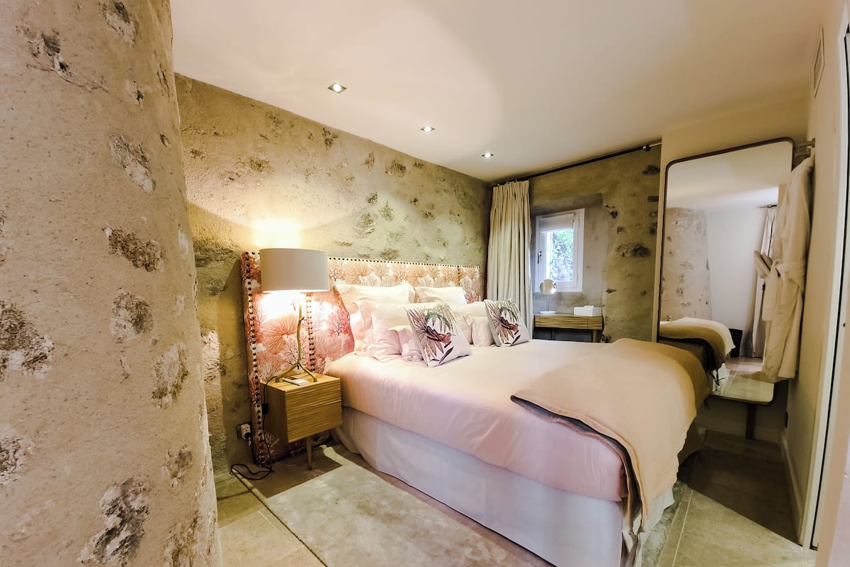 Une chambre à coucher avec un grand lit, une table de chevet avec une lampe, un miroir et une fenêtre avec des rideaux.