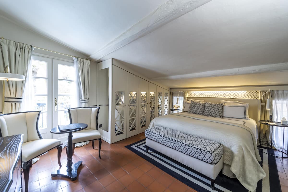 Une chambre d’hôtel avec un grand lit, un coin salon avec des fauteuils, une petite table et une vue sur l’extérieur par les fenêtres.