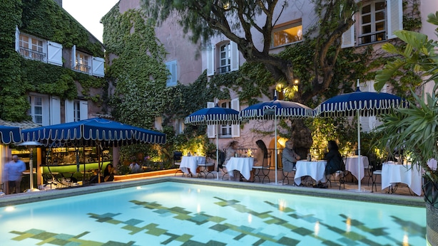 Una piscina con diseños de mosaicos, mesas al aire libre con sombrillas y un edificio con muros cubiertos de hiedra.