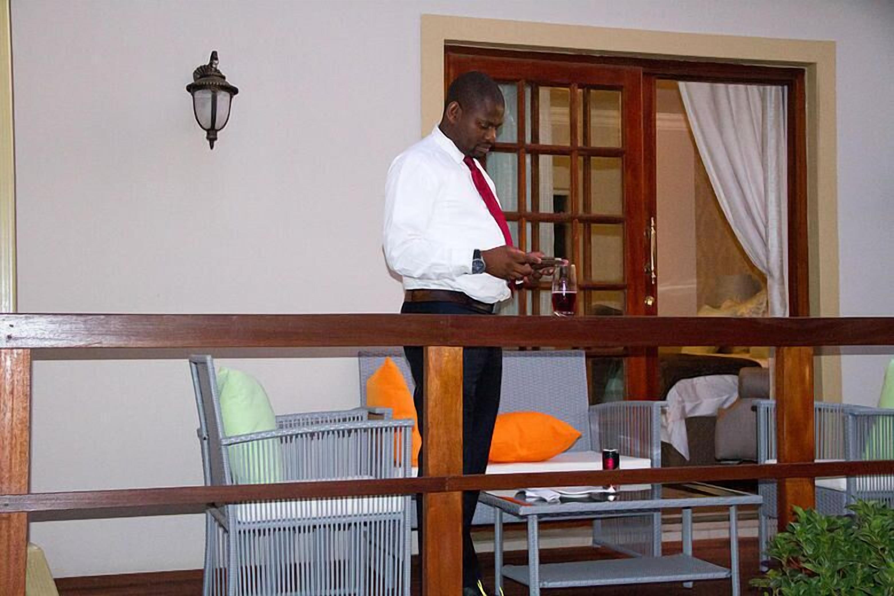 A man standing on a balcony with a view of a room with a bed, a table, and chairs.